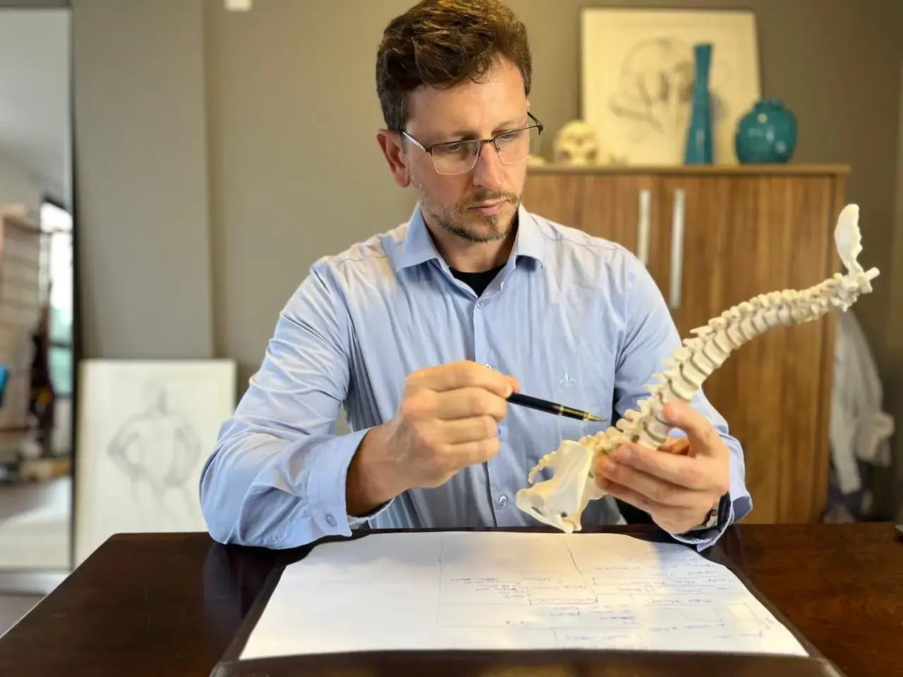 Leonardo Schneider, Fisioterapeuta e Osteopata em Florianópolis, explicando a coluna vertebral com um modelo, à mesa do consultório no Itacorubi
