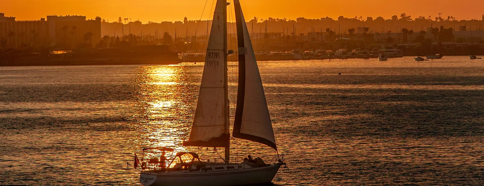 Veleiro navegando num mar calmo ao pôr do sol, com a cidade ao fundo