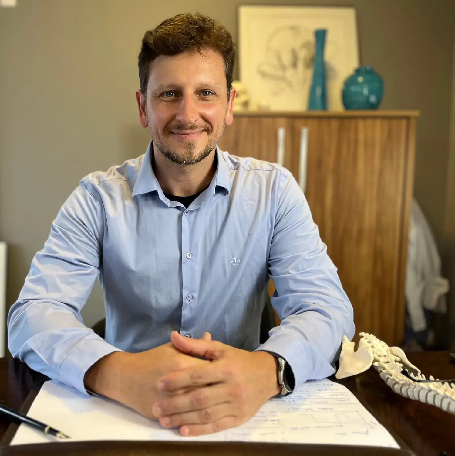 Leonardo Schneider sentado à mesa de madeira do consultório, com um modelo de coluna ao lado